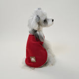 Small white dog wearing warm red fleece vest with white stitching, standing side view